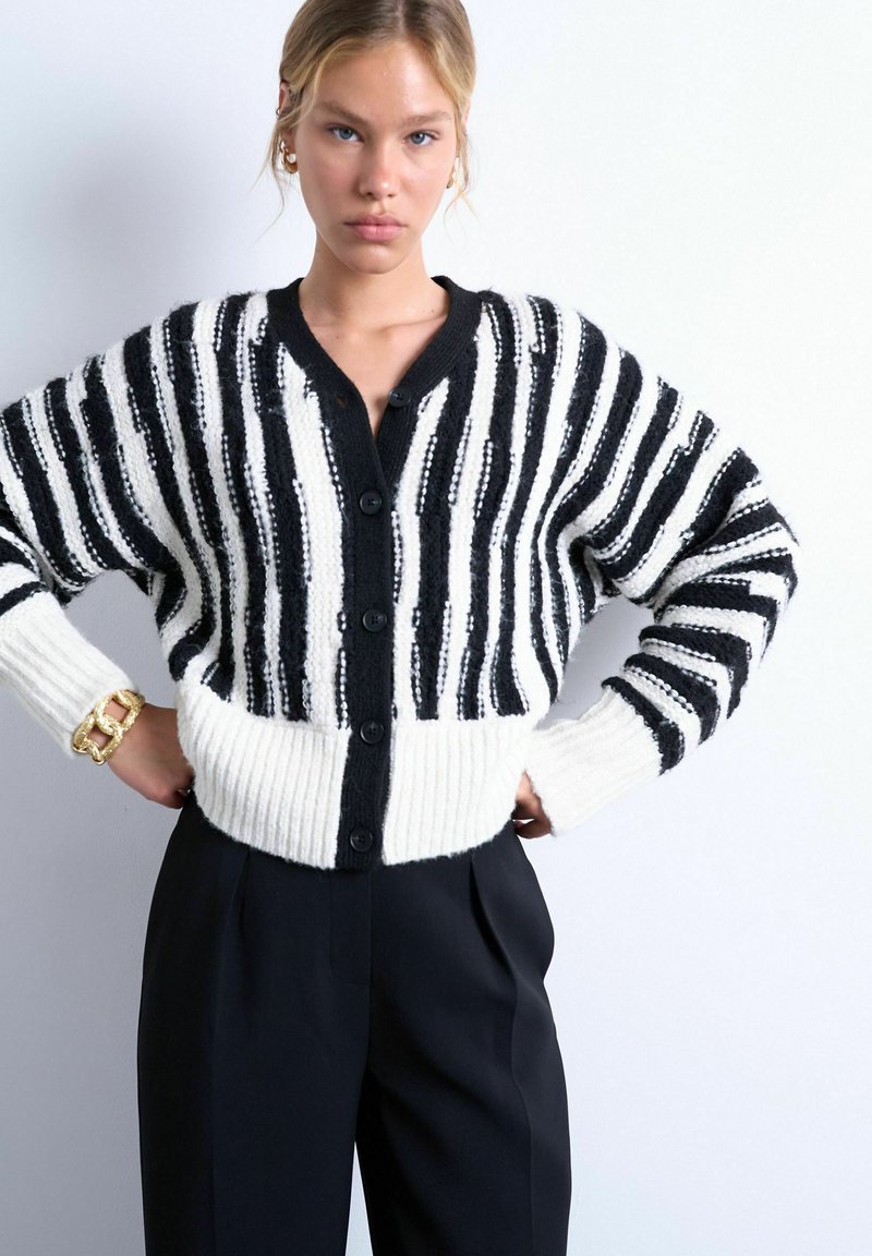 Jeune femme portant un cardigan boutonné à rayures noires et blanches et un pantalon taille haute noir, debout les mains sur les hanches sur fond blanc.