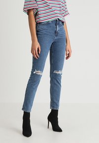 Jean en denim bleu taille haute avec des déchirures aux genoux, coupe slim, associés à des bottines à talons noires et un haut à rayures à manches courtes.