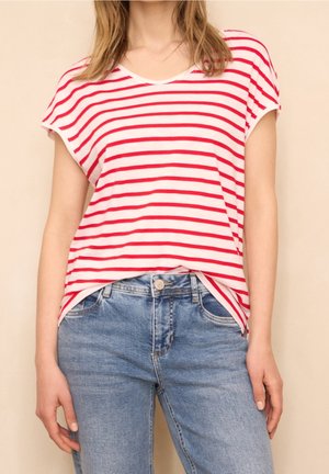 Mujer con una camisa blanca de manga corta con rayas horizontales rojas y vaqueros azules, de pie contra un fondo liso.