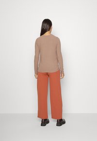 Blusa de manga longa em malha ribada de tom castanho claro, acompanhada por calças largas de cor ferrugem, vestidas com sapatos pretos de sola espessa.
