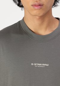 T-shirt gris foncé avec un col rond et des manches courtes. Présente un logo de marque blanc "G-STAR RAW" imprimé sur la poitrine. Texture lisse.