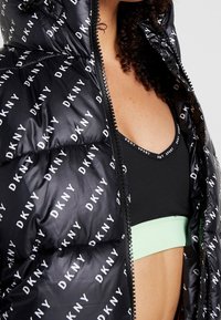 Černá péřová bunda s opakovaným bílým logem "DKNY", se zipovým otevíráním a velkou kapucí. Černá sportovní podprsenka s mátově zeleným lemováním pod ní.