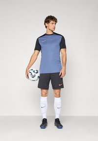 Nike sportsklær med en blå t-skjorte med sorte detaljer, grå shorts, hvite sokker og svarte fotballsko, som holder en fotball.