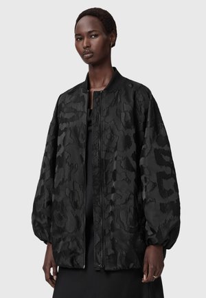 Dunkelhäutige Person mit kurzen Haaren, die eine schwarze übergroße, strukturierte Bomberjacke mit Reißverschluss und elastischen Bündchen trägt.