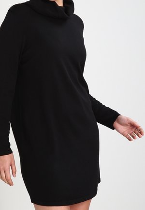 Kvinde iført en sort sweaterkjole med lange ærmer og løs rullekant mod en ensfarvet lys baggrund.