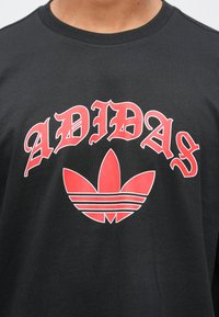 Melna kokvilnas T-krekls ar sarkanu "ADIDAS" uzrakstu un sarkanu trešlapju logotipu ar trim baltām svītrām. Īsas piedurknes un apaļa izgriezuma kakls.