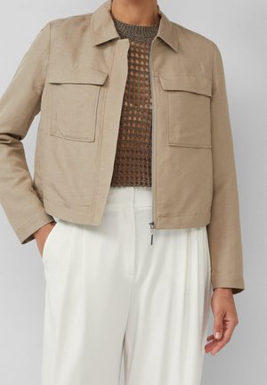 Veste courte beige avec poches avant sur un top marron en maille ouverte, associée à un pantalon blanc taille haute, main dans la poche.