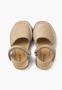 Beige nahkahiekat vauvoille suljettujen varpaiden, valkoisen ompelun ja tarranauhoilla varustettujen nilkkahihnojen kanssa valkoista taustaa vasten.