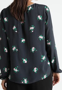 Blusa a maniche lunghe grigio scuro con piccoli motivi floreali rosa e foglie verdi. Tessuto liscio con una leggera lucentezza e accenti strutturati sottili.