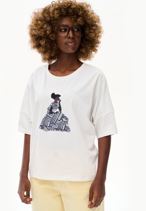 Kobieta z kręconymi włosami i okularami, ubrana w białą koszulkę z grafiką siedzącej postaci w wzorzystym niebiesko-białym ubraniu.