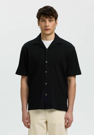 Mladý muž s krátkymi kučeravými vlasmi, ktorý má oblečenú čiernu krátku košeľu na gombíky cez biele tričko a béžové nohavice, stojaci pred jednoduchým pozadím.