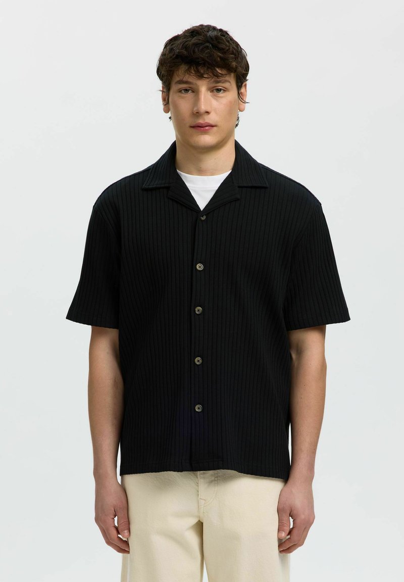 Jeune homme aux cheveux courts et bouclés portant une chemise noire à manches courtes boutonnée sur un T-shirt blanc et un pantalon beige, debout devant un fond uni.