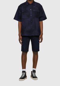 Camisa de manga corta de color navy con dos bolsillos frontales, cierre de botones y un diseño de panel geométrico. Combinada con pantalones cortos de mezclilla oscura y zapatillas altas negras.
