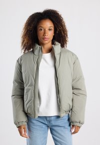 Chaqueta acolchada en verde claro, con cierre de cremallera frontal, cuello alto, puños y dobladillo elásticos, combinada con una camiseta blanca y unos vaqueros azules.