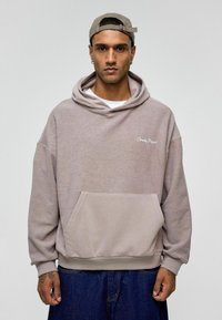 Oversized beige hoodie met een voorzak, ribbelboorden en -zoombaan, voorzien van de tekst "Eternity Project" in het wit geborduurd op de borst.