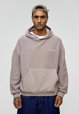 Oversized beige hoodie met een voorzak, ribbelboorden en -zoombaan, voorzien van de tekst "Eternity Project" in het wit geborduurd op de borst.