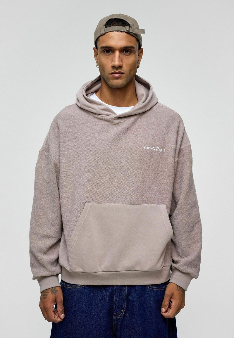 Oversized beige hoodie met een voorzak, ribbelboorden en -zoombaan, voorzien van de tekst "Eternity Project" in het wit geborduurd op de borst.