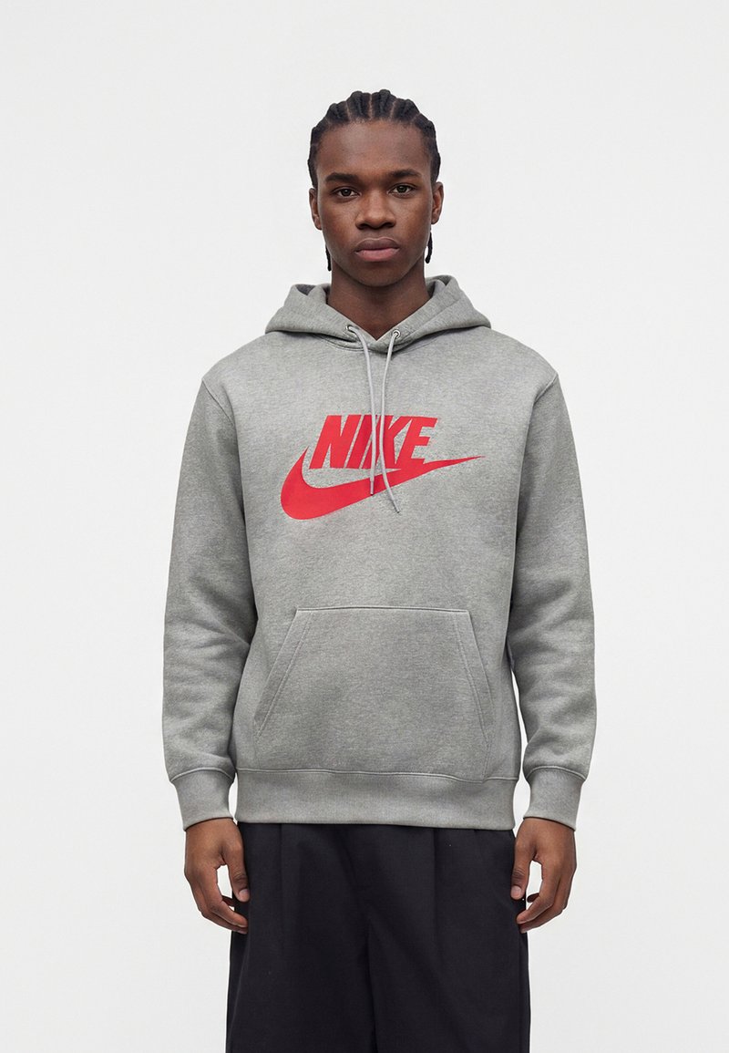 Joven con trenzas, vestido con una sudadera gris con capucha con el logo rojo de Nike y pantalones negros, de pie frente a un fondo blanco liso.