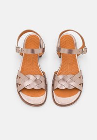 Sandalias metálicas de color rosa dorado con correas trenzadas, hebilla en el tobillo y plantilla texturizada en tono marrón. Diseño plano adecuado para uso casual.