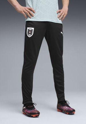Persona vistiendo pantalones de fútbol negros de Puma con el logo de la Asociación de Fútbol de Austria y zapatillas negras y rojas, con las manos en las caderas, de pie sobre un suelo gris.