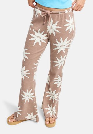 Femme portant un pantalon beige à jambes larges avec de grands motifs solaires blancs et une taille élastiquée avec cordon, associé à des sandales camel et un top bleu.