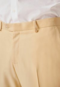 Beige maßgeschneiderte Hose mit glatter Textur, featuring vorderen Falten, einer Knopfverschluss und Gürtelschlaufen am Bund.