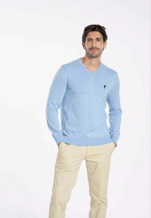 Pull bleu clair en V en tissu doux, avec des poignets et un ourlet côtelés. Présente un petit logo sur la poitrine gauche. Associé à un pantalon beige.