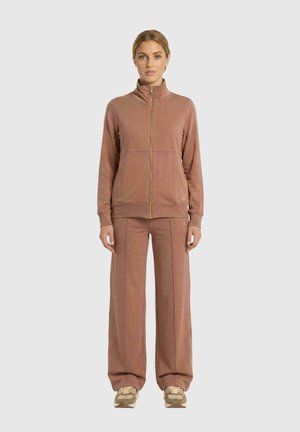 Le mannequin se tient devant un fond uni, vêtu d’une veste zippée rose poudré, d’un pantalon assorti à jambes larges et de baskets beiges.