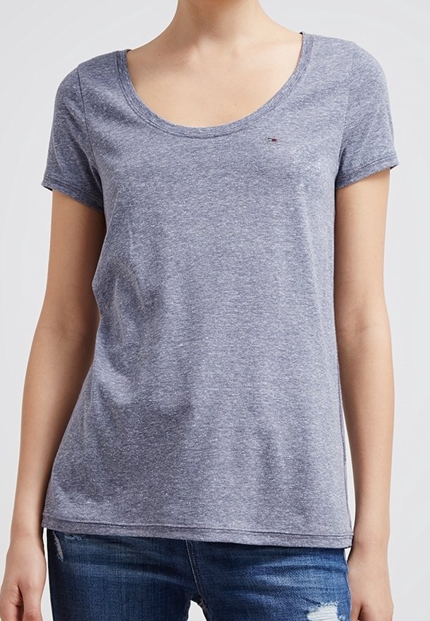 Kvinde iført en ensfarvet, gråmeleret t-shirt med rund hals og korte ærmer samt slidte blå jeans, stående mod en lys baggrund.