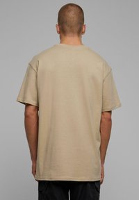 Beige kokvilnas T-krekls ar brīvu piegriezumu, īsām piedurknēm un standarta apaļu kakla izgriezumu. Audums izskatās mīksts ar gludu tekstūru.