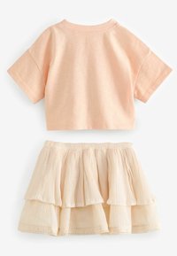 Pfirsichfarbene, kurzärmelige Baumwoll-Crop-Top, kombiniert mit einem mehrlagigen, cremefarbenen Rock mit Falten und einer Spitzenverzierung am Saum.