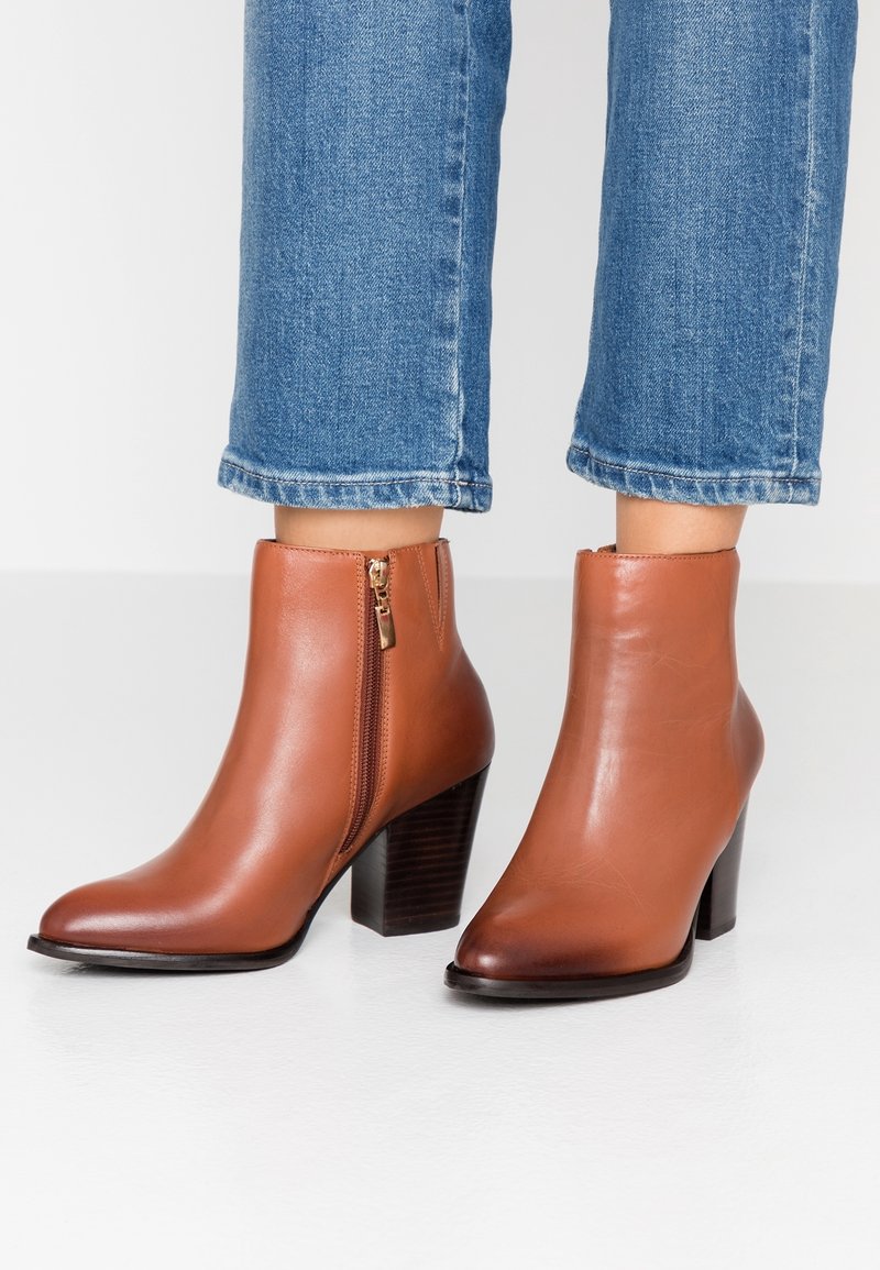 Bottines en cuir marron avec fermetures éclair latérales et talons carrés portées avec un jean denim bleu court sur une surface blanche.