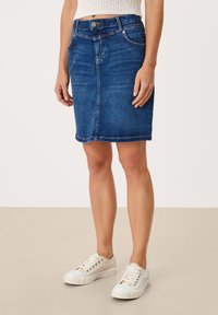 Falda vaquera de color azul medio, hasta la rodilla con corte recto. Cuenta con bolsillos delanteros y un ligero desgaste, combinada con zapatillas blancas.