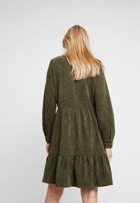 Femme blonde vue de dos portant une robe mini à manches longues, de couleur vert olive, à volants avec des poignets boutonnés, sur fond uni.