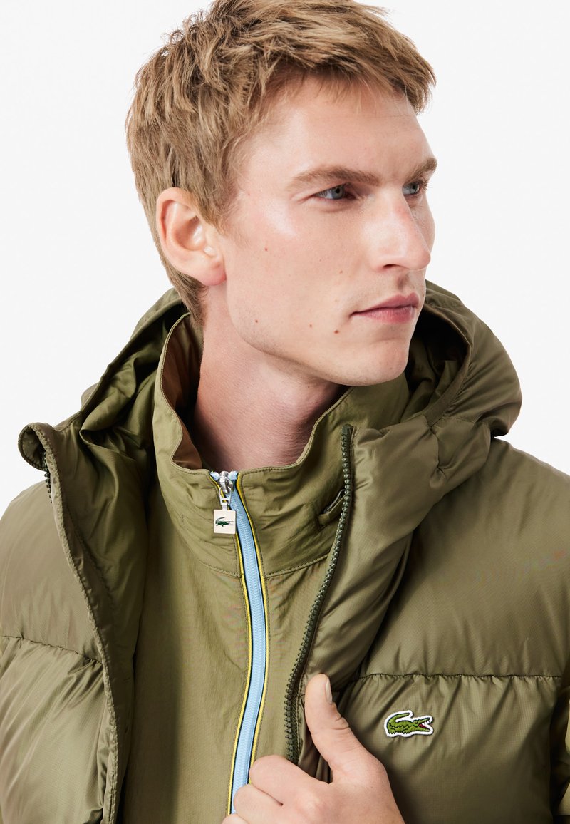 Chaqueta de plumas verde oliva con capucha, que presenta un cierre de cremallera completo y acentos en azul. Incluye un pequeño logotipo bordado en el pecho. Textura suave.