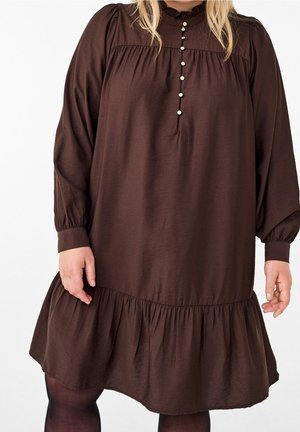 Losvallende donkerbruine jurk met lange mouwen, parelachtige knopen aan de voorkant en een gerimpelde zoom, gedragen met zwarte panty's.