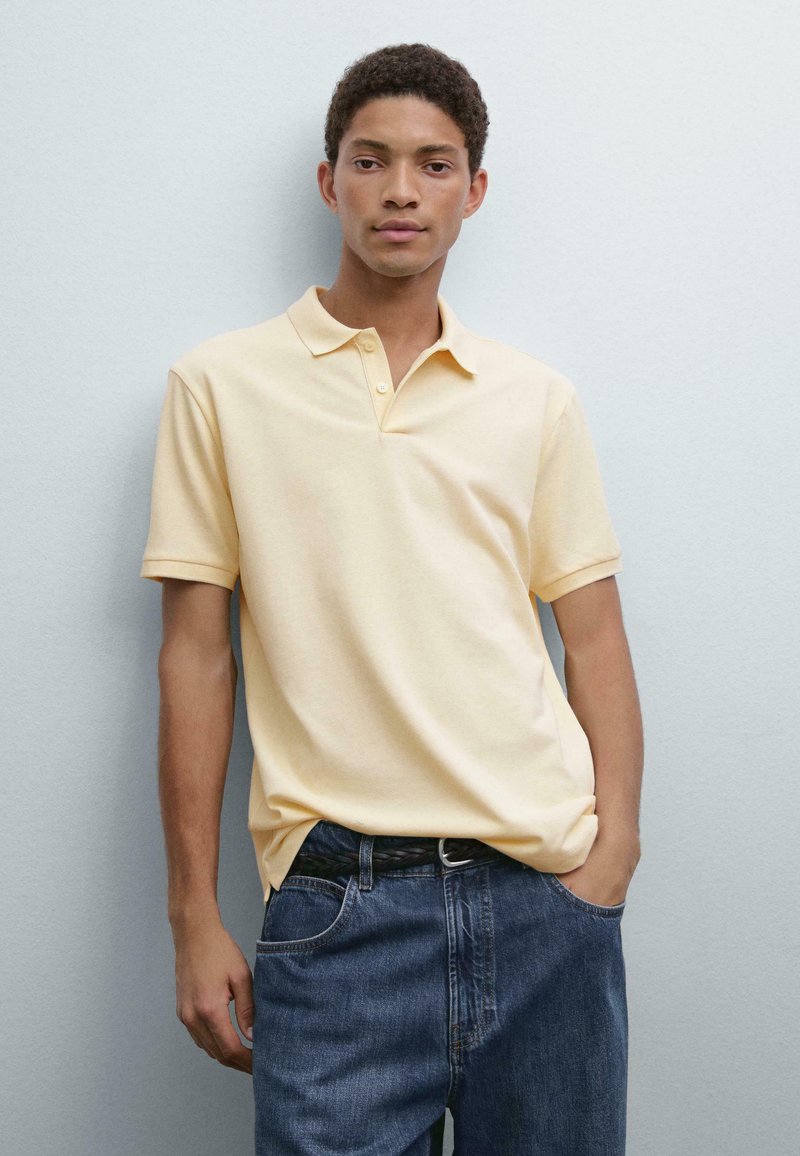 Polo in morbido cotone giallo chiaro, maniche corte, con pattina a tre bottoni, abbinato a jeans in denim blu larghi e una cintura nera.