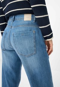 Jeans in denim blu con vita alta, due tasche posteriori e un'etichetta del marchio ben visibile. Il tessuto presenta una texture slavata con dettagli di cucitura sottili.