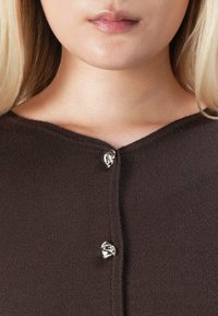 Close-up van de onderkant van het gezicht en de nek van een vrouw die een donkerbruin gebreid vest draagt met twee glanzende, onregelmatige zilveren knopen vastgemaakt.