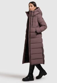 Palton lung, căptușit, de culoare mauve, cu guler înalt, buzunare cu fermoar și fermoar pe toată lungimea. Are un design texturat, capitonat și o glugă largă.