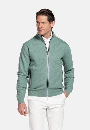 Man draagt een lichtgroene jas met rits over een wit shirt en witte broek, staat met één hand in de zak en kijkt opzij.