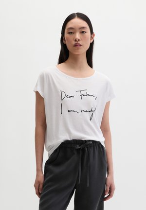 Jeune femme aux longs cheveux foncés portant un T-shirt blanc où il est écrit « Cher futur, je suis prête ! » et un pantalon noir à cordon.