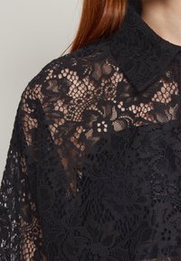Chemise en dentelle florale noire avec un col présentant des motifs transparents complexes sur l'épaule et le haut de la poitrine, portée par une personne aux longs cheveux roux.