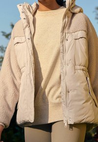 Persoon in een beige gevlekte trui, crèmekleurige sherpa fleece jas met een geborduurde voorkant en ribbelige beige broek, buiten in natuurlijk licht.
