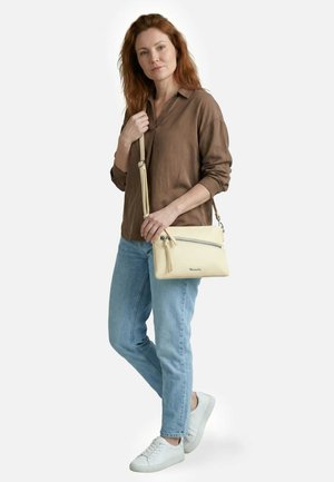 Vrouw in bruin shirt en lichtblauwe spijkerbroek staat met een crèmekleurige schoudertas met ritsdetail, draagt witte sneakers tegen een witte achtergrond.