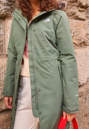 Personne portant un manteau d'hiver vert North Face avec une capuche bordée de fourrure, un pantalon beige et tenant un sac rouge devant un mur en pierre.