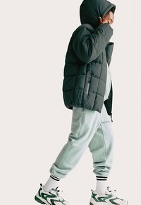 Černá péřová bunda s kapucí, šedé tepláky a tenisky se zelenými detaily. Oblečení má prošívanou texturu a sportovní design.