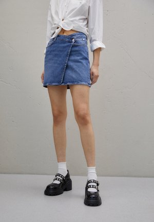 Personne portant une mini-jupe en denim bleu, une chemise blanche retroussée et nouée à la taille, des chaussettes blanches et des chaussures compensées Mary Jane noires avec des brides.