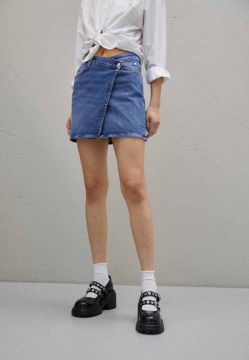 Personne portant une mini-jupe en denim bleu, une chemise blanche retroussée et nouée à la taille, des chaussettes blanches et des chaussures compensées Mary Jane noires avec des brides.