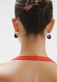 Pendientes de aro de oro con un acento circular azul marino, llevados por una persona con un peinado de moño y una blusa de tirantes roja.